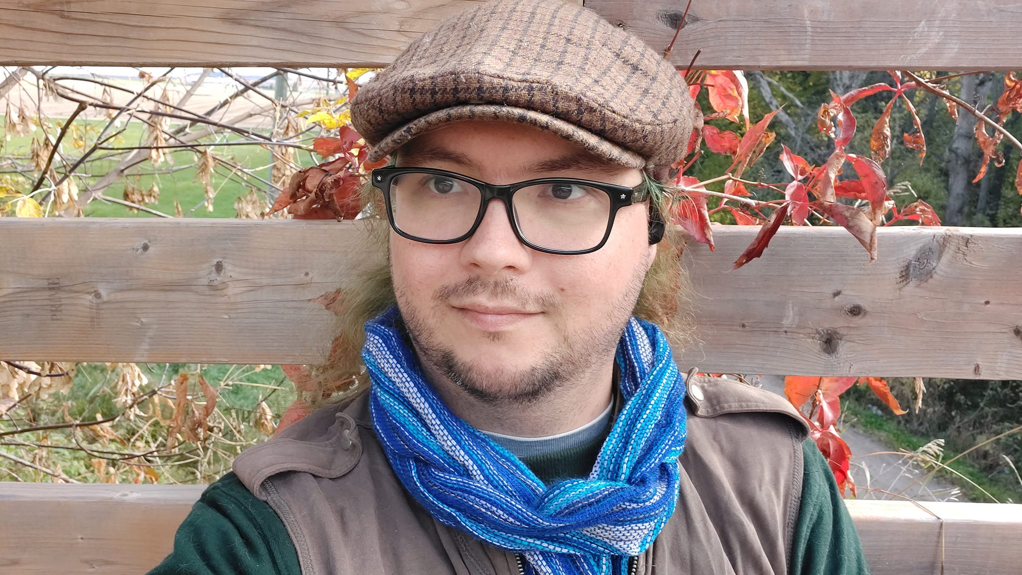 A man with long green hair wearing a green long sleave shirt with a brown zippered sweater vest, a blue scarf and a brown flat cap sits leaning against the wodden safety bars on a bridge 